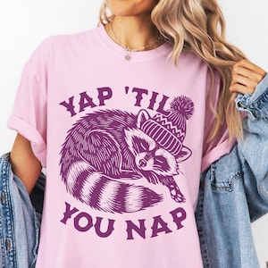 May include: A pink t-shirt with a purple graphic of a raccoon wearing a winter hat and the text "YAP 'TIL YOU NAP".