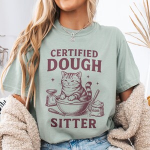 May include: A light green t-shirt with the text "CERTIFIED DOUGH SITTER" and a graphic of a cat in a bowl with baking tools. The shirt is a soft, comfortable material.