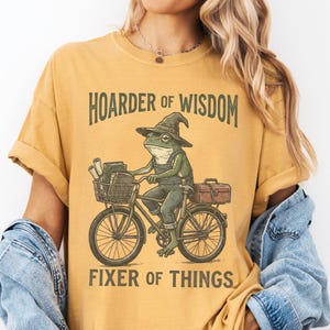 May include: Mustard-colored t-shirt featuring a frog riding a bicycle with a toolbox and basket. The frog wears overalls and a witch hat. Text on the shirt reads "HOARDER OF WISDOM" and "FIXER OF THINGS."