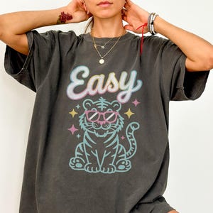 K&ouml;nnte beinhalten: Dunkelgraues T-Shirt mit dem Wort "Easy" in einer Pastellschrift &uuml;ber einem Zeichentricktiger mit Sonnenbrille. Der Tiger ist blau und t&uuml;rkis umrandet, mit rosa und gelben Akzenten. Das Shirt hat kurze &Auml;rmel.