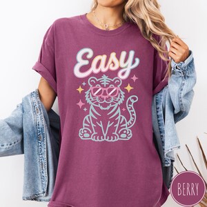 K&ouml;nnte beinhalten: Beerenfarbenes T-Shirt mit dem Wort "Easy" in einer verspielten Schriftart &uuml;ber einem Zeichentricktiger mit Sonnenbrille. Der Tiger ist hellblau mit rosa Akzenten und von Sternen umgeben. Das Shirt hat hochgekrempelte &Auml;rmel.