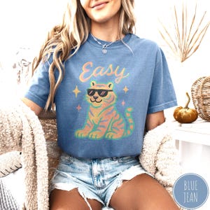May include: Blue jean-colored t-shirt with the word "Easy" in a pastel font above a cartoon tiger wearing sunglasses. The tiger is orange and pink with black stripes. The shirt has a relaxed fit.