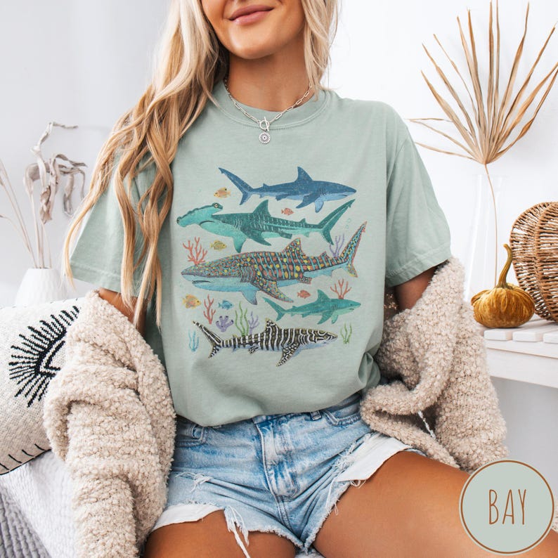 May include: A light green t-shirt featuring a watercolor illustration of various shark species, including hammerhead, whale, and tiger sharks. The design includes coral and small fish, creating an ocean-themed graphic.