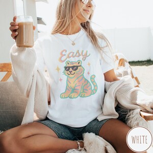 May include: White t-shirt with a colorful tiger graphic wearing sunglasses and the word "Easy" in pastel colors. The tiger design includes orange, pink, and yellow hues. The shirt is a casual style.