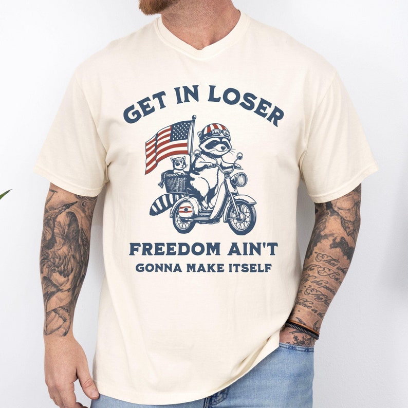 May include: A cream colored t-shirt with a vintage style graphic of a raccoon riding a scooter with an American flag in the background. The text "Get in Loser, Freedom Ain't Gonna Make Itself" is printed on the shirt.