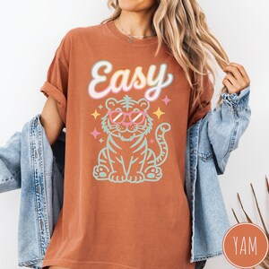 K&ouml;nnte beinhalten: Rostfarbenes T-Shirt mit dem Wort "Easy" in einer Pastellschrift &uuml;ber einem Zeichentricktiger mit Sonnenbrille. Der Tiger ist t&uuml;rkis und rosa umrandet, mit Sternakzenten.