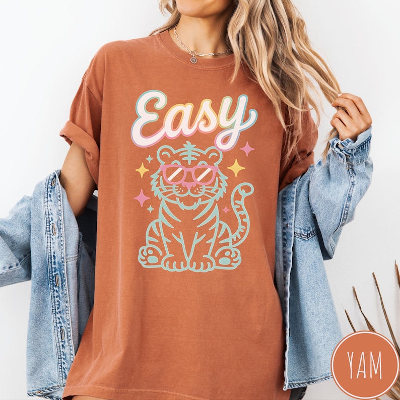 K&ouml;nnte beinhalten: Rostfarbenes T-Shirt mit dem Wort "Easy" in einer Pastellschrift &uuml;ber einem Zeichentricktiger mit Sonnenbrille. Der Tiger ist t&uuml;rkis und rosa umrandet, mit Sternakzenten.