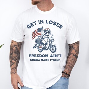 May include: White t-shirt with a blue and red American flag, a raccoon riding a scooter, and the text "Get in loser, freedom ain't gonna make itself".