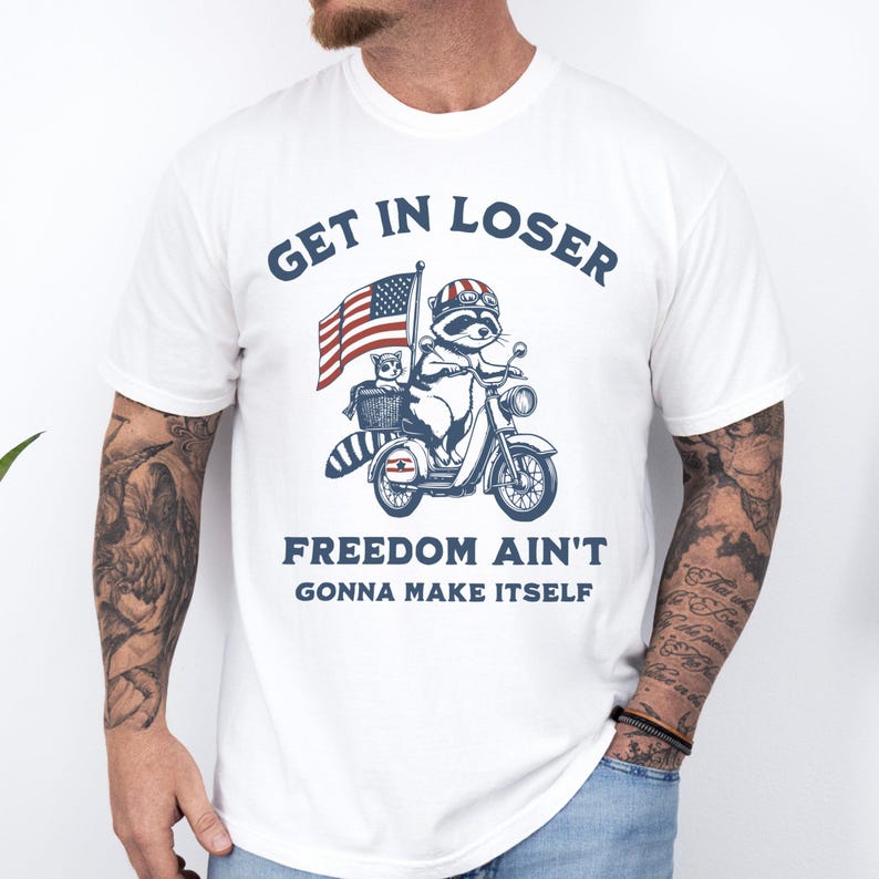 May include: White t-shirt with a blue and red American flag, a raccoon riding a scooter, and the text "Get in loser, freedom ain't gonna make itself".