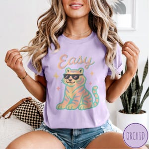 May include: A lilac t-shirt featuring a cartoon tiger wearing sunglasses, with the word "Easy" above it. The tiger has orange and yellow stripes and is surrounded by small star graphics. The shirt is a casual wear item.