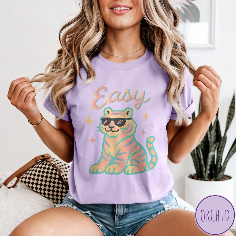 May include: A lilac t-shirt featuring a cartoon tiger wearing sunglasses, with the word "Easy" above it. The tiger has orange and yellow stripes and is surrounded by small star graphics. The shirt is a casual wear item.