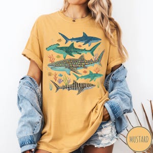 May include: Mustard-colored t-shirt featuring a colorful illustration of various shark species, including hammerhead and whale sharks, with coral and fish accents. The shirt has a relaxed fit and rolled sleeves.