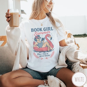 May include: White t-shirt with a graphic that reads "BOOK GIRL Summer" above an illustration of a woman reading in a flamingo float. The text "Dive into a good book!" is below the illustration.