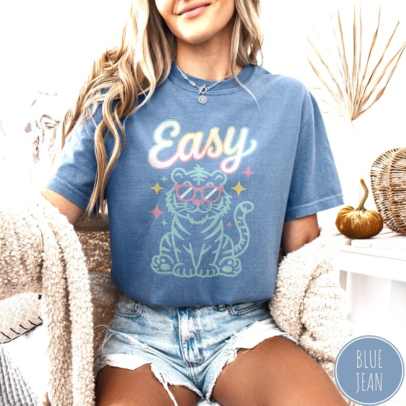 K&ouml;nnte beinhalten: Blaues kurz&auml;rmeliges T-Shirt mit dem Wort "Easy" in Pastellschrift &uuml;ber einem Zeichentricktiger mit Sonnenbrille. Der Tiger ist hellblau mit rosa und gelben Akzenten. Das Shirt hat eine blaue Farbe.