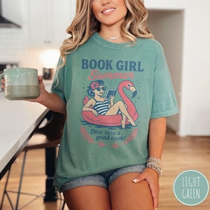 May include: Light green t-shirt with a graphic that reads "BOOK GIRL Summer" above an illustration of a woman reading in a flamingo floatie. The text "Dive into a good book!" is below the illustration. The shirt is paired with denim shorts.