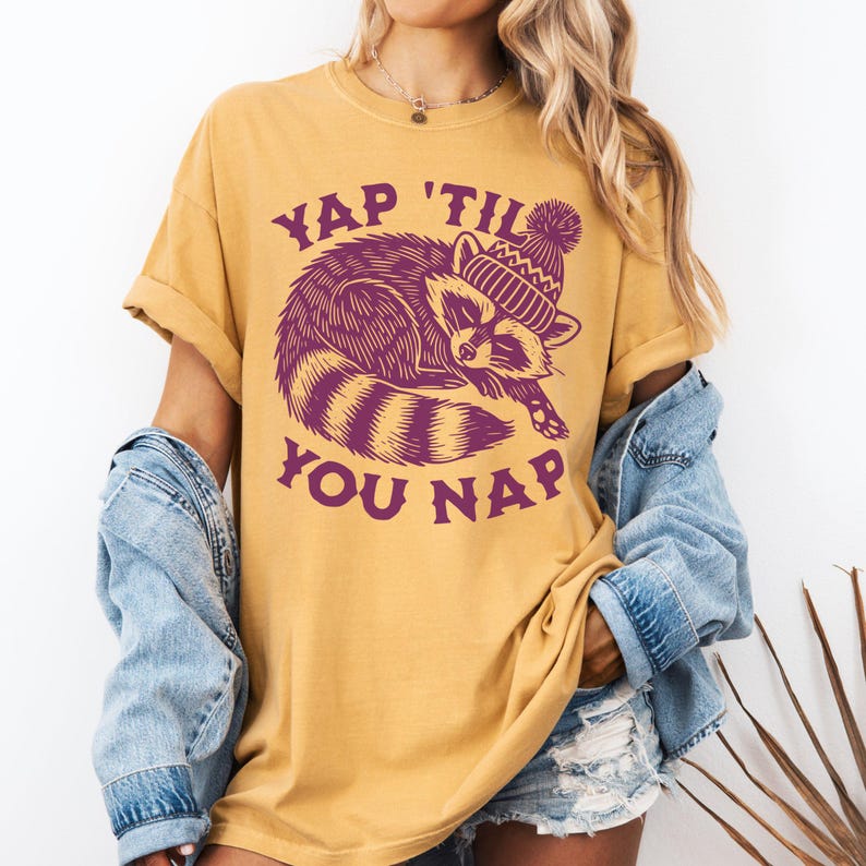 May include: A mustard yellow t-shirt with a purple raccoon wearing a purple and white knit hat. The text "YAP 'TIL YOU NAP" is printed on the shirt.