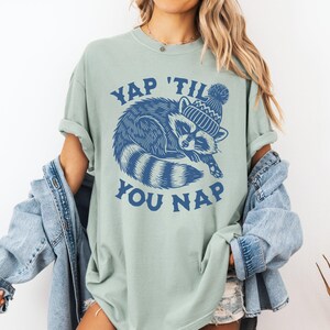May include: A light green t-shirt with a blue graphic of a raccoon wearing a winter hat and the text "YAP 'TIL YOU NAP".