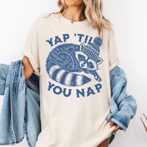 May include: A white t-shirt with a blue graphic of a raccoon wearing a blue and white hat. The text "YAP 'TIL YOU NAP" is printed in blue below the raccoon.