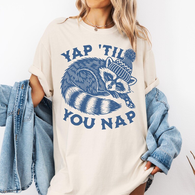 May include: A white t-shirt with a blue graphic of a raccoon wearing a blue and white hat. The text "YAP 'TIL YOU NAP" is printed in blue below the raccoon.