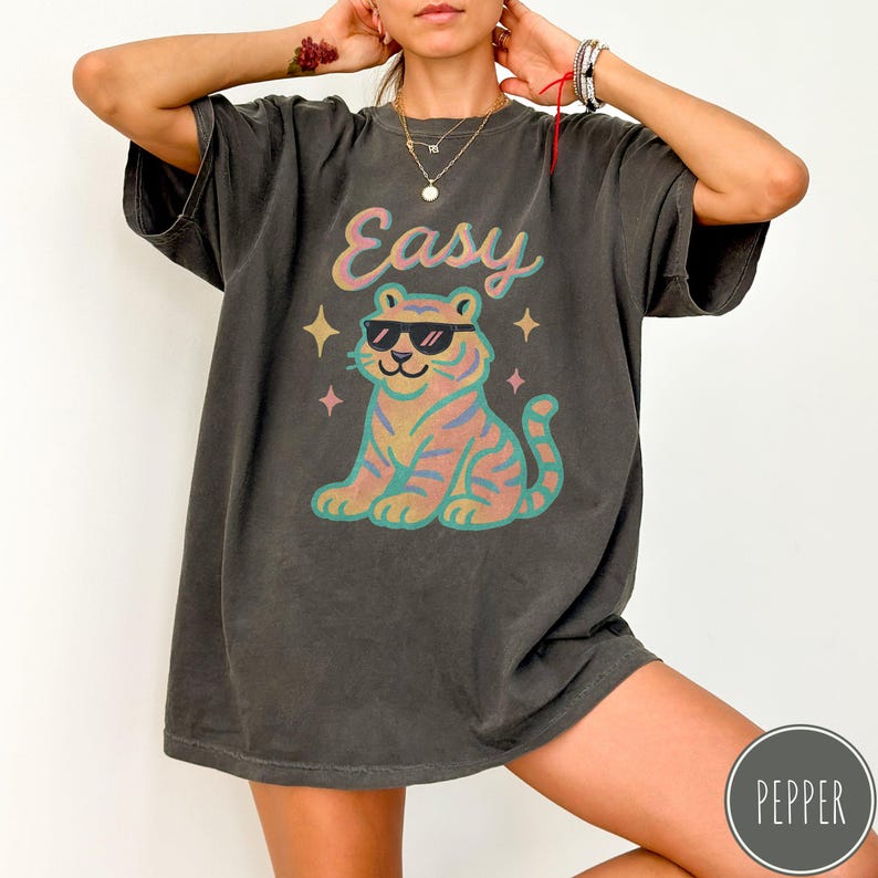 May include: Dark grey t-shirt with the word "Easy" in a script font above a colorful tiger wearing sunglasses. The tiger is in shades of pink, blue, and yellow. The shirt has a relaxed fit.