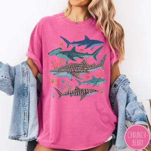 May include: A hot pink t-shirt featuring a colorful illustration of various shark species, including hammerhead, whale, and zebra sharks. The design includes coral and fish. The text "CHRUNCH-BERRY" is in a circle on the bottom right.