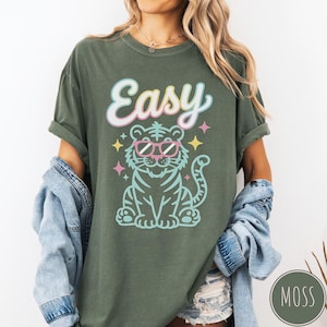 K&ouml;nnte beinhalten: Olivgr&uuml;nes T-Shirt mit dem Wort "Easy" in Schreibschrift &uuml;ber einem Zeichentricktiger mit rosa Sonnenbrille. Der Tiger ist hellblau mit schwarzen Streifen und von bunten Sternen umgeben.