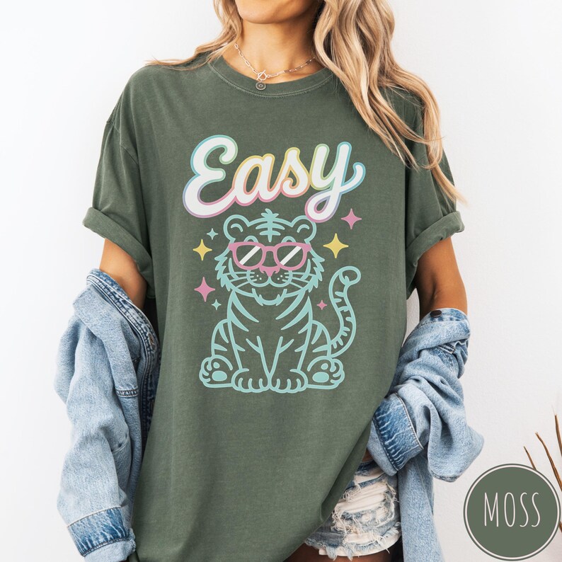 K&ouml;nnte beinhalten: Olivgr&uuml;nes T-Shirt mit dem Wort "Easy" in Schreibschrift &uuml;ber einem Zeichentricktiger mit rosa Sonnenbrille. Der Tiger ist hellblau mit schwarzen Streifen und von bunten Sternen umgeben.