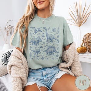May include: Sage green t-shirt featuring a blue dinosaur illustration. The design includes various dinosaur species, plants, and a spiral fossil. The shirt is a comfortable, casual style, perfect for dinosaur enthusiasts.