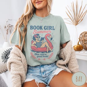 May include: A light green t-shirt with the text "BOOK GIRL Summer" and an illustration of a woman reading in a flamingo float. The text "Dive into a good book!" is also included. The shirt is a casual summer style.