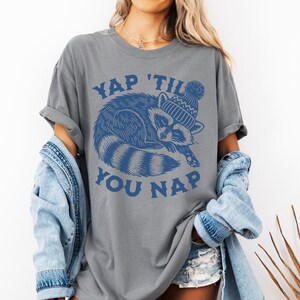 May include: Gray t-shirt with a blue graphic of a raccoon wearing a winter hat and the text "YAP 'TIL YOU NAP".