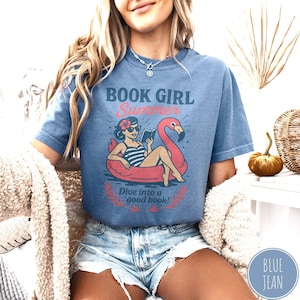 May include: A blue short-sleeved t-shirt with the words "BOOK GIRL Summer" and a graphic of a woman reading in a flamingo float. The text "Dive into a good book!" is below the graphic. The shirt is worn with denim shorts.