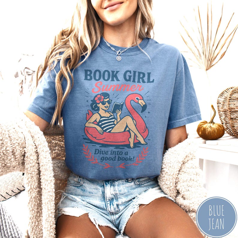 May include: A blue short-sleeved t-shirt with the words "BOOK GIRL Summer" and a graphic of a woman reading in a flamingo float. The text "Dive into a good book!" is below the graphic. The shirt is worn with denim shorts.