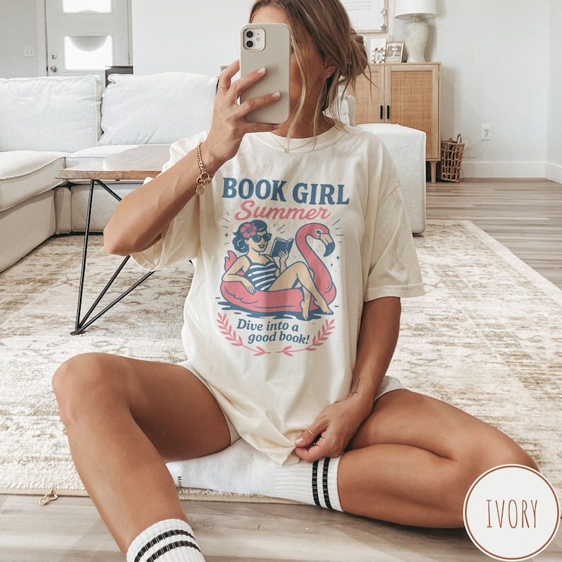 May include: Ivory-colored t-shirt with the text "BOOK GIRL Summer" and an illustration of a woman reading in a flamingo float. The shirt also has the text "Dive into a good book!"