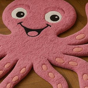 May include: A pink octopus-shaped rug with a textured surface. The rug features a smiling face with large white eyes, black pupils, and a black mouth. The eight tentacles are decorated with light pink oval suction cup patterns throughout the design.