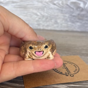 May include: A small, hand-sculpted clay frog figurine with a rounded, tan and brown mottled body. The frog has two black eyes and an open mouth revealing a bright pink tongue. It is positioned above a brown card with the text Handcrafted Just For You.