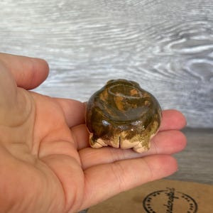 May include: A small, rounded ceramic frog figurine with a glossy, mottled olive-green and brown glaze. The sculpture features a smooth, curved back and visible hind legs tucked underneath, showcasing a handcrafted, earthy aesthetic.