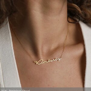 以下が含まれることがあります： 筆記体で「Kimberly」と書かれたゴールドカラーのネックレス。繊細なチェーンに吊り下げられています。画像は、人の首に着用されているネックレスを示しています。ネックレスは316Lステンレス鋼製です。