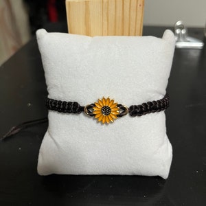 May include: A black braided bracelet with a gold sunflower charm. The sunflower is yellow with a black center.