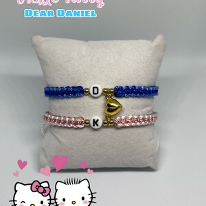 May include: Two macrame bracelets, one blue and one pink, each with a letter charm and a gold heart charm. The bracelets are displayed on a white pillow. The image includes the text "Hello Kitty Dear Daniel" and a Hello Kitty graphic.