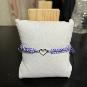 May include: A purple friendship bracelet with a silver heart charm. The bracelet is displayed on a white velvet cushion.