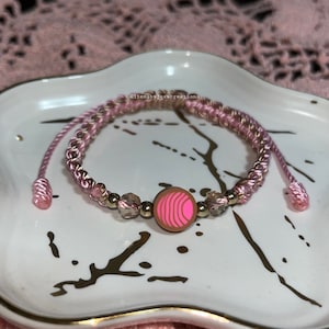 May include: Pink and gold braided bracelet with a pink and brown donut charm. The bracelet is on a white plate with a gold rim.