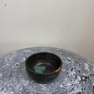 May include: A small, round ceramic bowl with a dark, matte black exterior and a smooth, deep interior. The inside of the bowl features a prominent splash of turquoise glaze at the base and a single, distinct golden-brown accent on the rim.