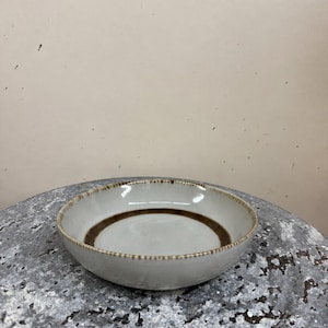 May include: A shallow, round ceramic bowl with a light gray, speckled glaze. The interior features a prominent, dark brown circular band painted near the base, and the outer rim is decorated with a series of small, evenly spaced brown dashes.
