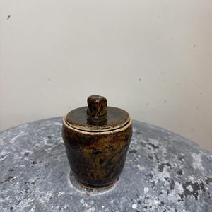 May include: A small, cylindrical ceramic jar with a dark brown, mottled glaze and a matching lid. The lid features a small, raised, knob-like handle at the center. The surface of the jar has a textured, earthy appearance with variations in color.