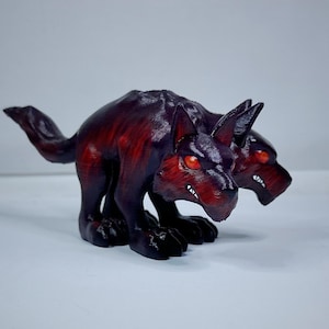 May include: A two-headed Cerberus figurine, a mythical dog from Greek mythology. The sculpture is primarily dark red and black, with glowing red eyes and sharp white teeth. The dog is in a crouching position.