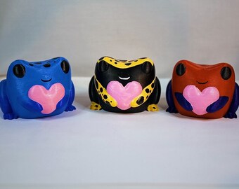 May include: Three small frog figurines, each holding a pink heart. The frogs are painted in different colors: blue, black with yellow spots, and red.