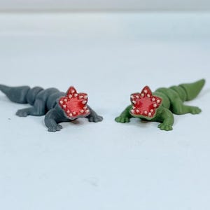 May include: Two small, 3D printed, alligator-like figures with open mouths. One is gray and the other is green. Both have red, star-shaped mouths with white teeth.