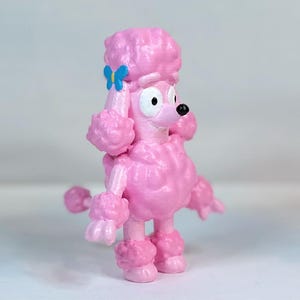 K&ouml;nnte beinhalten: Eine rosafarbene Pudelfigur mit einer blauen Schleife in Form eines Schmetterlings auf dem Kopf. Die Figur hat einen Comic-Stil mit gro&szlig;en Augen und einem breiten L&auml;cheln.