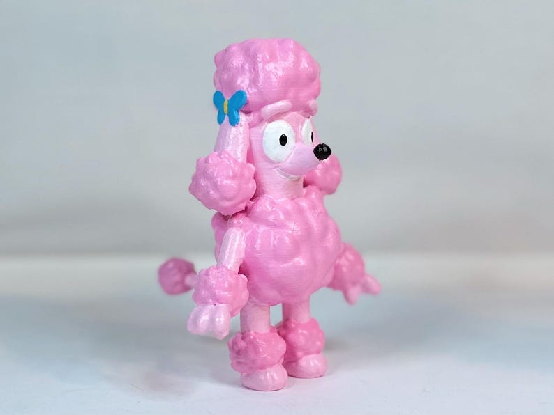 K&ouml;nnte beinhalten: Eine rosafarbene Pudelfigur mit einer blauen Schleife in Form eines Schmetterlings auf dem Kopf. Die Figur hat einen Comic-Stil mit gro&szlig;en Augen und einem breiten L&auml;cheln.
