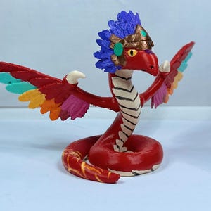 May include: A red and white snake with a colorful feathered headdress and wings. The snake is coiled and has a white and black striped pattern on its body. The headdress is made of blue, green, and gold feathers.